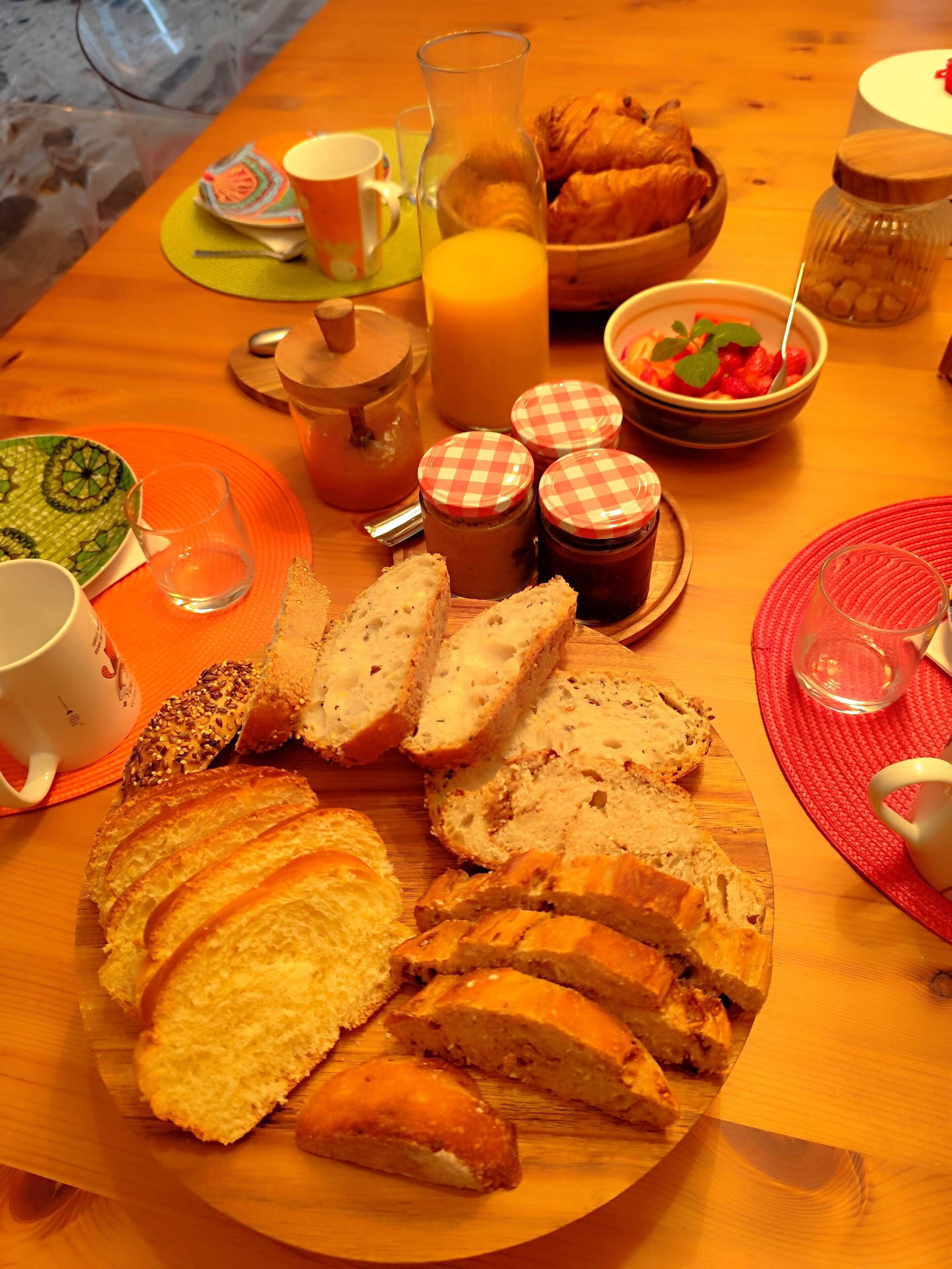 Ein Holztisch mit Brot, einem Croissant, einer Obstschale, einer Kerze und einem Glas Orangensaft.