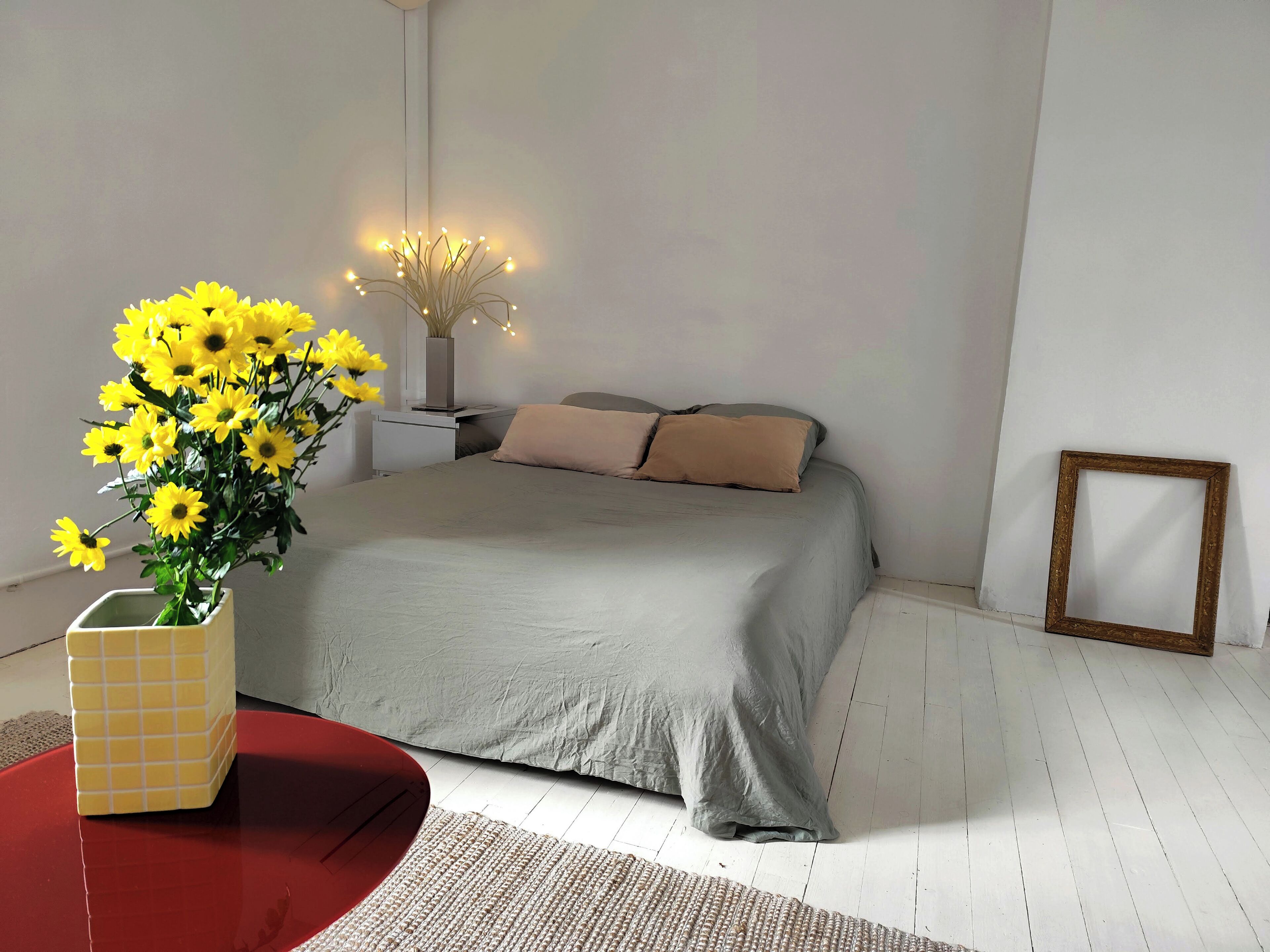 A bedroom with a bed, a nightstand, a vase of yellow flowers, a red table, and a picture frame on the wall.