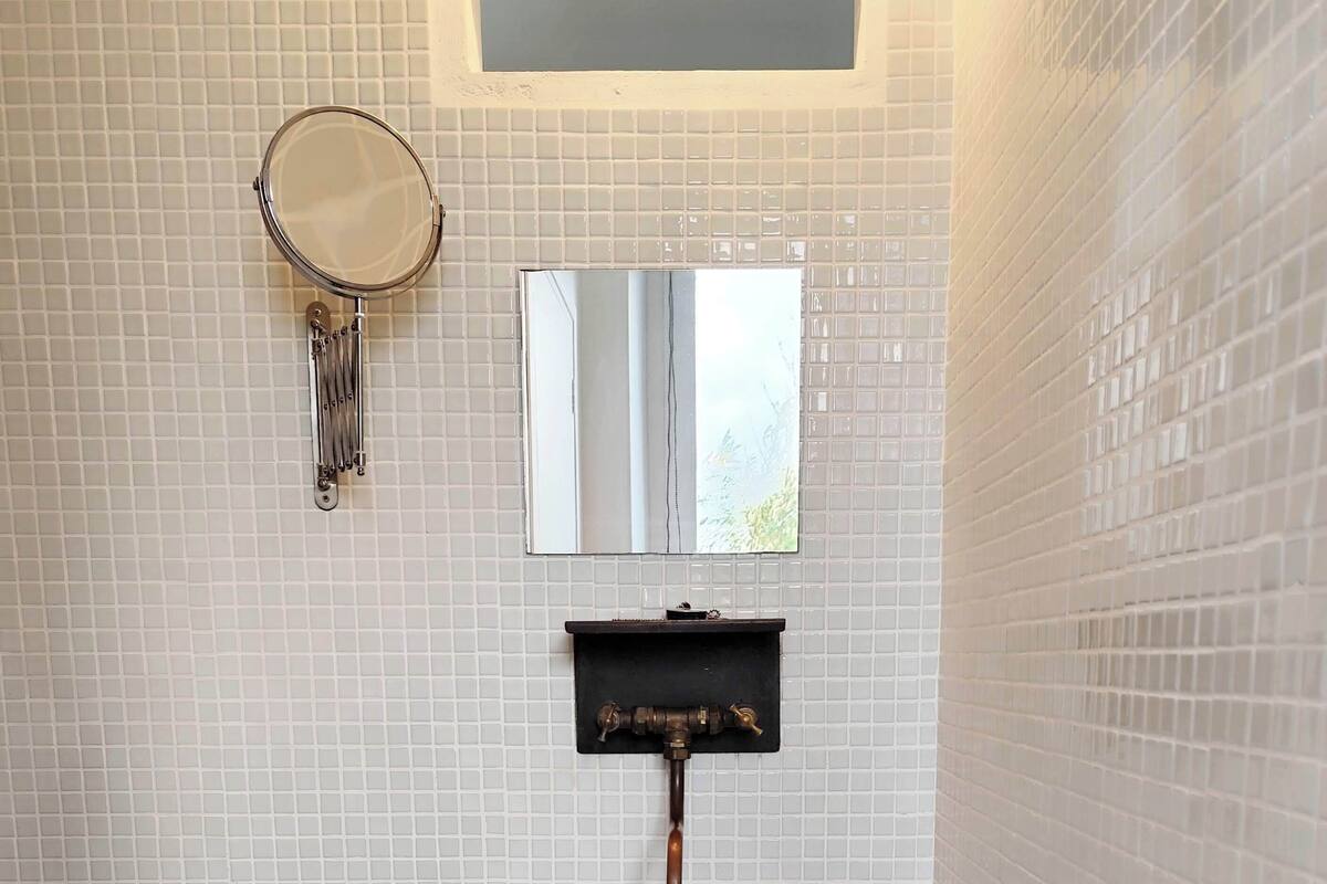 A bathroom with a white tiled wall, a round mirror, a rectangular sink with a black stool, and a white trash bin.
