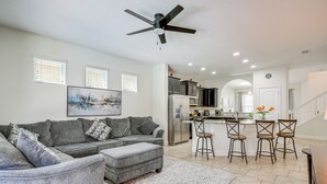 A spacious living room with a grey sectional sofa, a ceiling fan, and a kitchen area with a bar stool set.