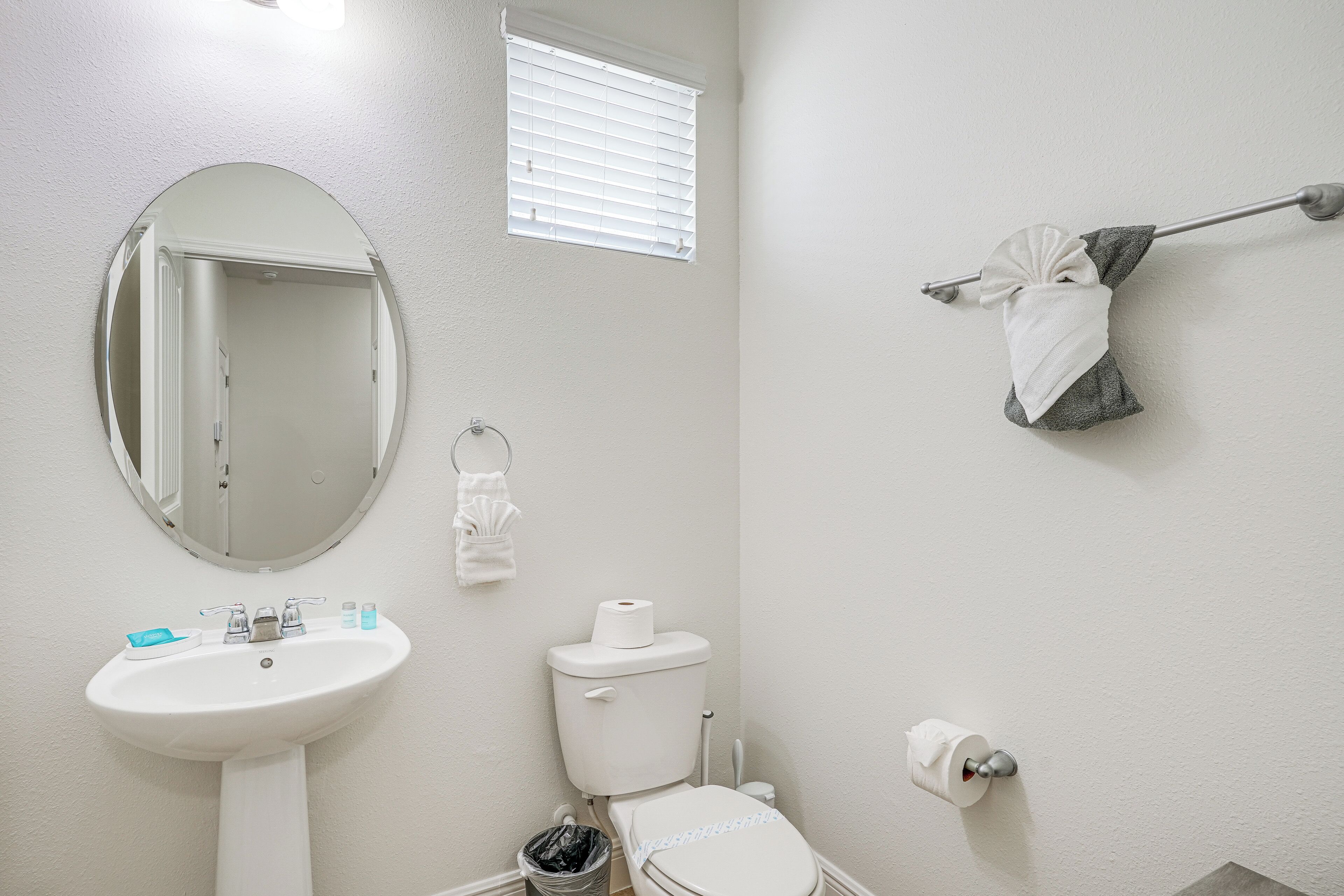 Ein Badezimmer mit weißem Waschbecken, Spiegel, Toilette, Handtuchhalter und einem Fenster mit Jalousien.