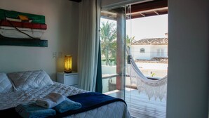 Un dormitorio con una cama, una mesita de noche con lámpara, una hamaca en el balcón y vistas del edificio y palmeras.