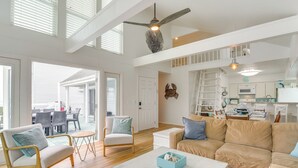 Un vaste salon doté d’un haut plafond, d’un plancher en bois et d’un grand canapé beige. On y trouve un coin repas avec une table et des chaises, ainsi qu’une cuisine équipée de placards et d’un comptoir.