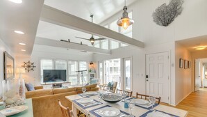 Un coin repas avec une longue table dressée pour six personnes, un ventilateur de plafond et une grande fenêtre équipée de stores.