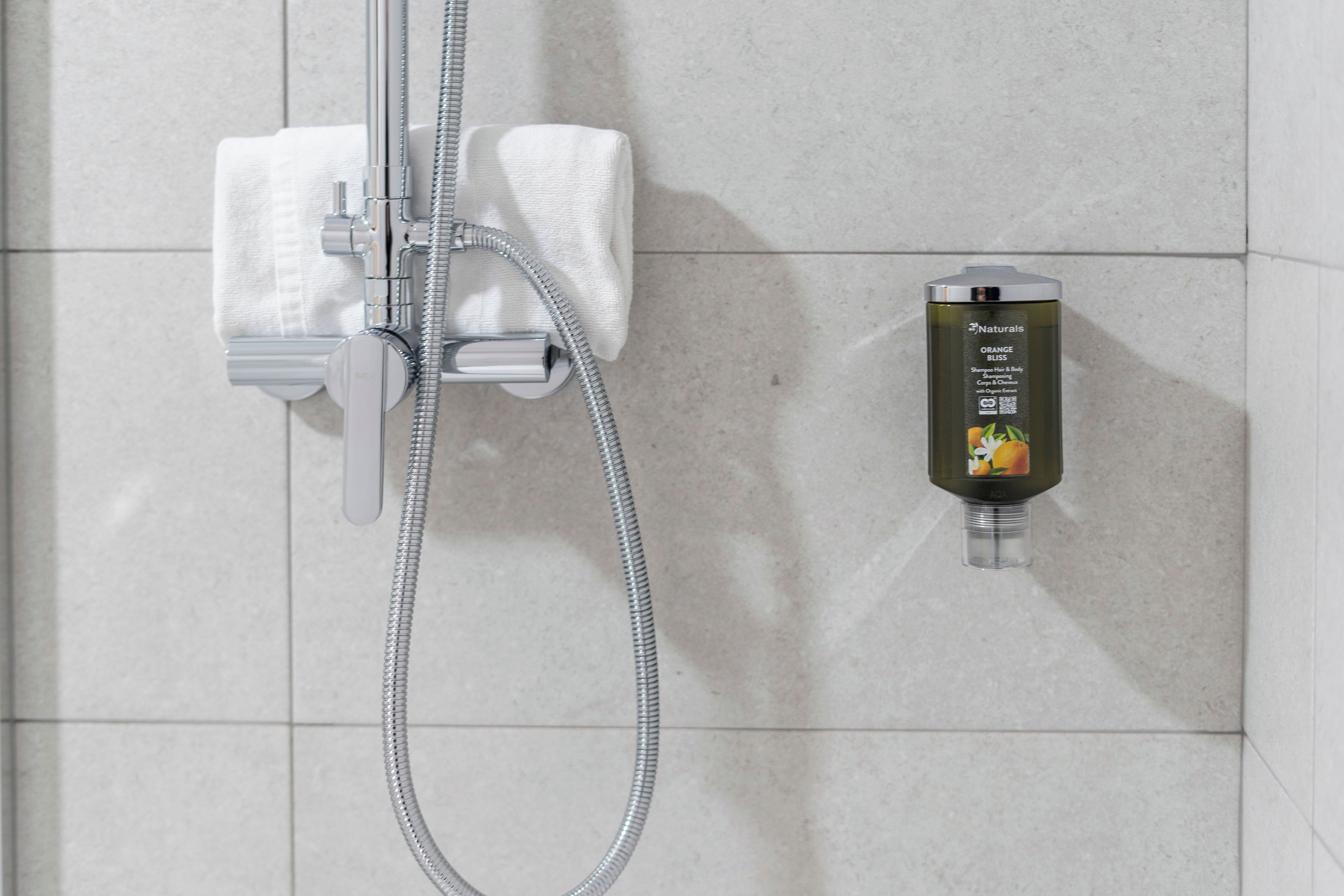 Chuveiro com mangueira manual e um frasco de shampoo de flor de laranjeira na parede.