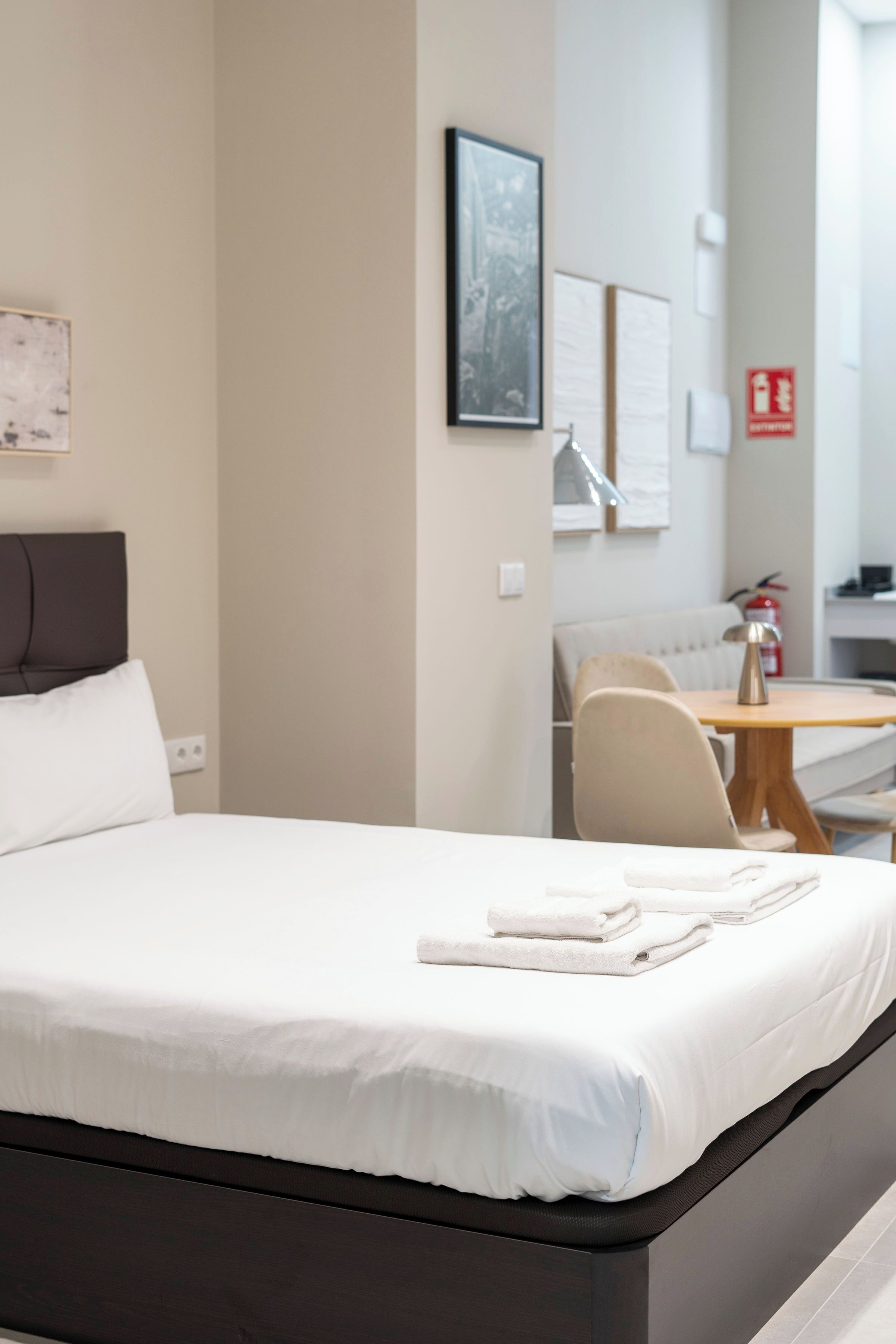 A neatly made bed with white linens, a small wooden table with a lamp, and a chair.