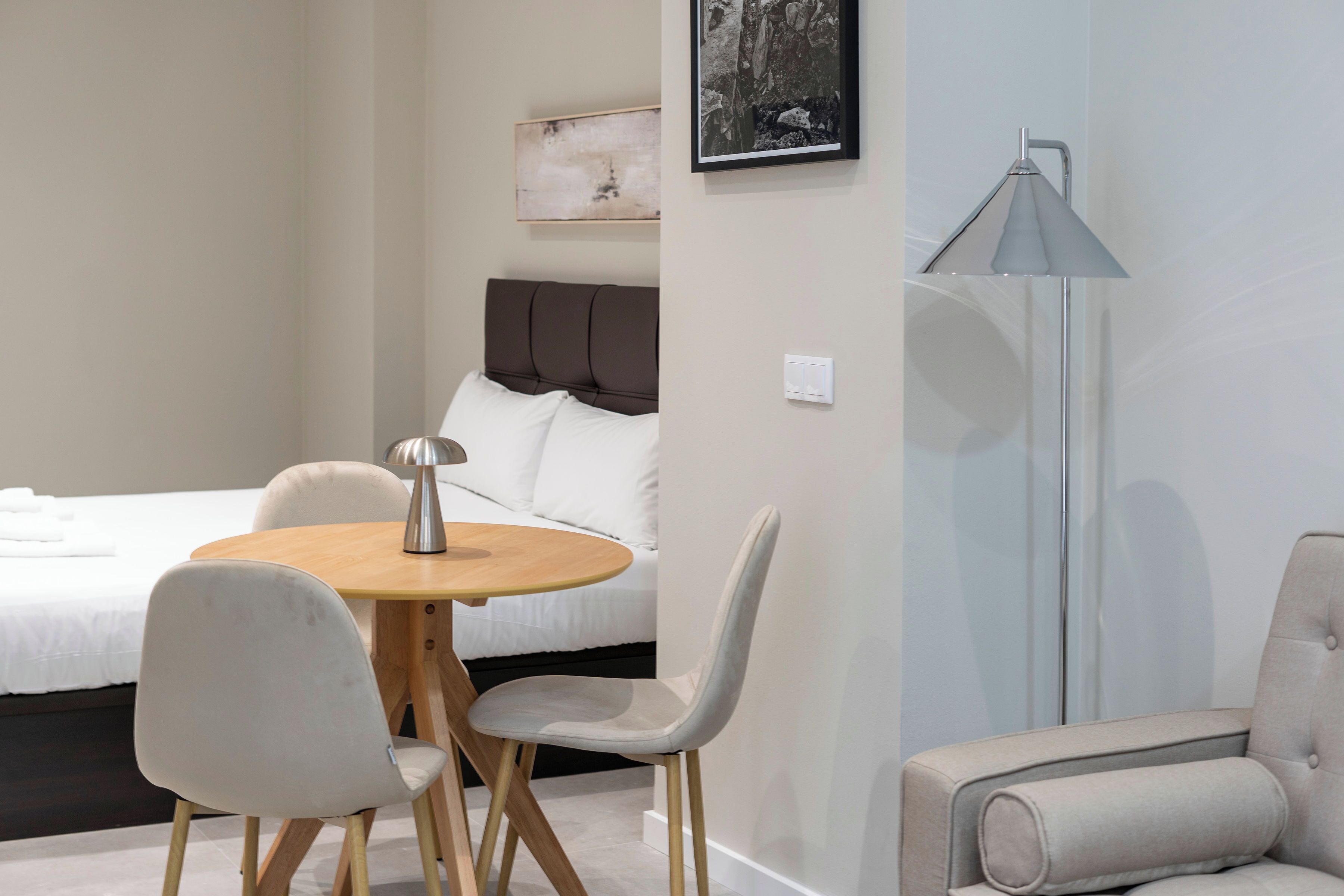 A modern hotel room with a bed, a round wooden table with two chairs, a floor lamp, and a large painting on the wall.
