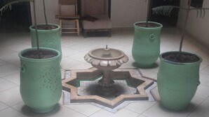A tiled floor with four green planters, a central decorative piece, and a wooden chair in a room with a door and a window in the background. - Hôtel de la paix (Marrakech)