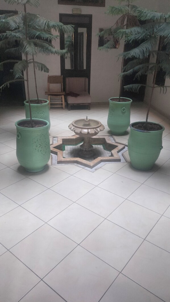 Un sol carrelé agrémenté de quatre jardinières vertes, une pièce décorative centrale et une chaise en bois, dans une pièce avec une porte et une fenêtre en arrière-plan.