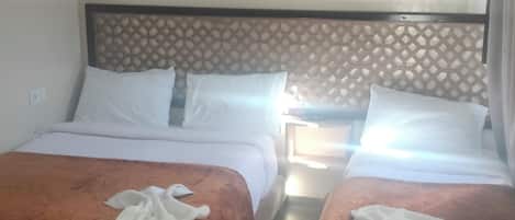A hotel room with two beds, each with a brown bedspread and white pillows. There is a wooden nightstand between the beds with a lamp. The room has a simple, modern design with a plain wall and a ceiling with recessed lighting.