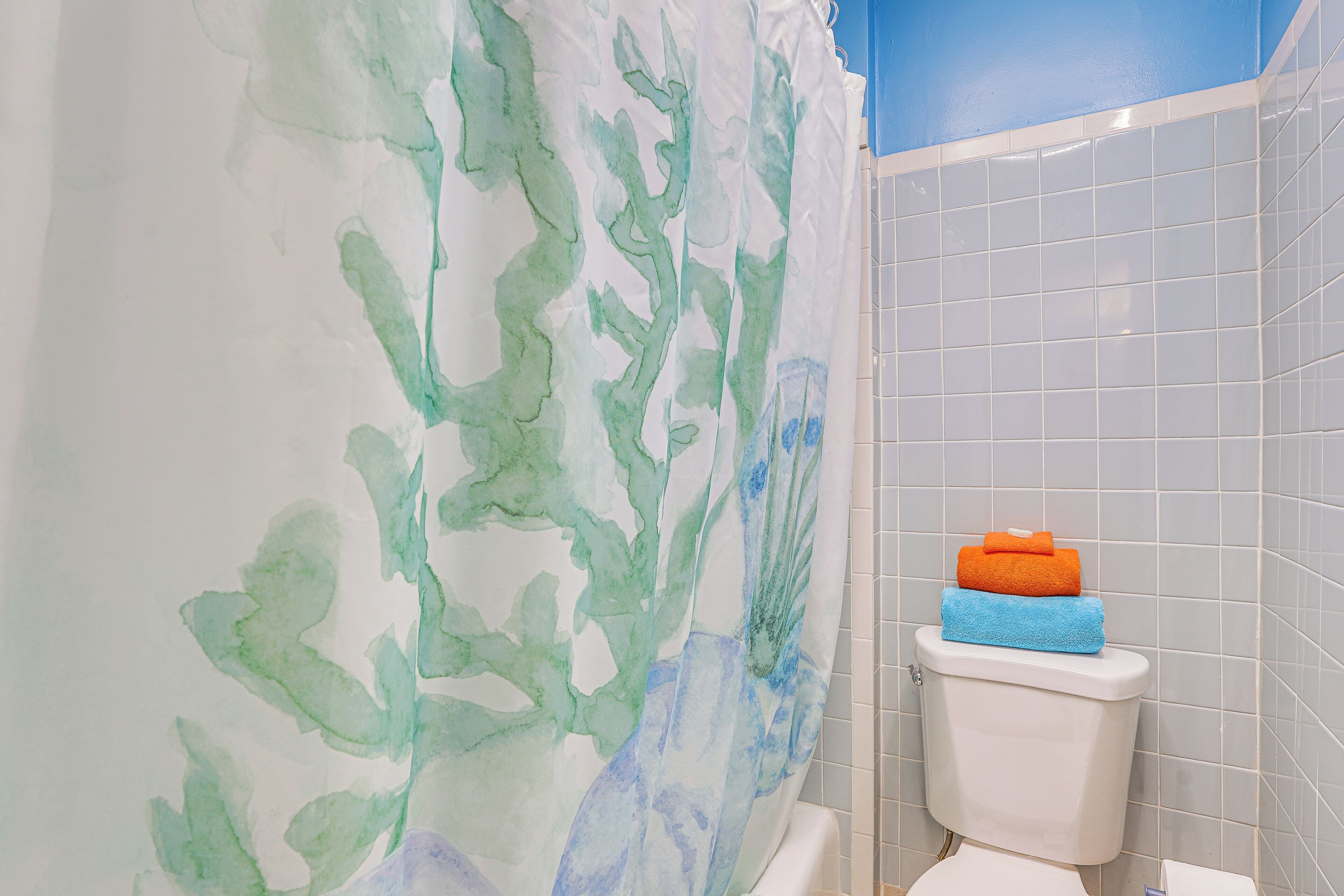 A bathroom with a white toilet, a blue and orange towel on the tank, and a wall with a green and blue abstract design.