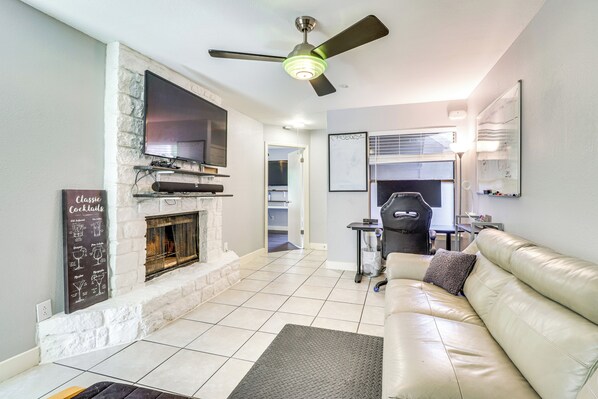 A living room with a fireplace, a flat-screen TV mounted above it, a leather sofa, and a ceiling fan. - 4 Mi to Dtwn: Gem w/ Pool Access & Patio in Austin (Austin)