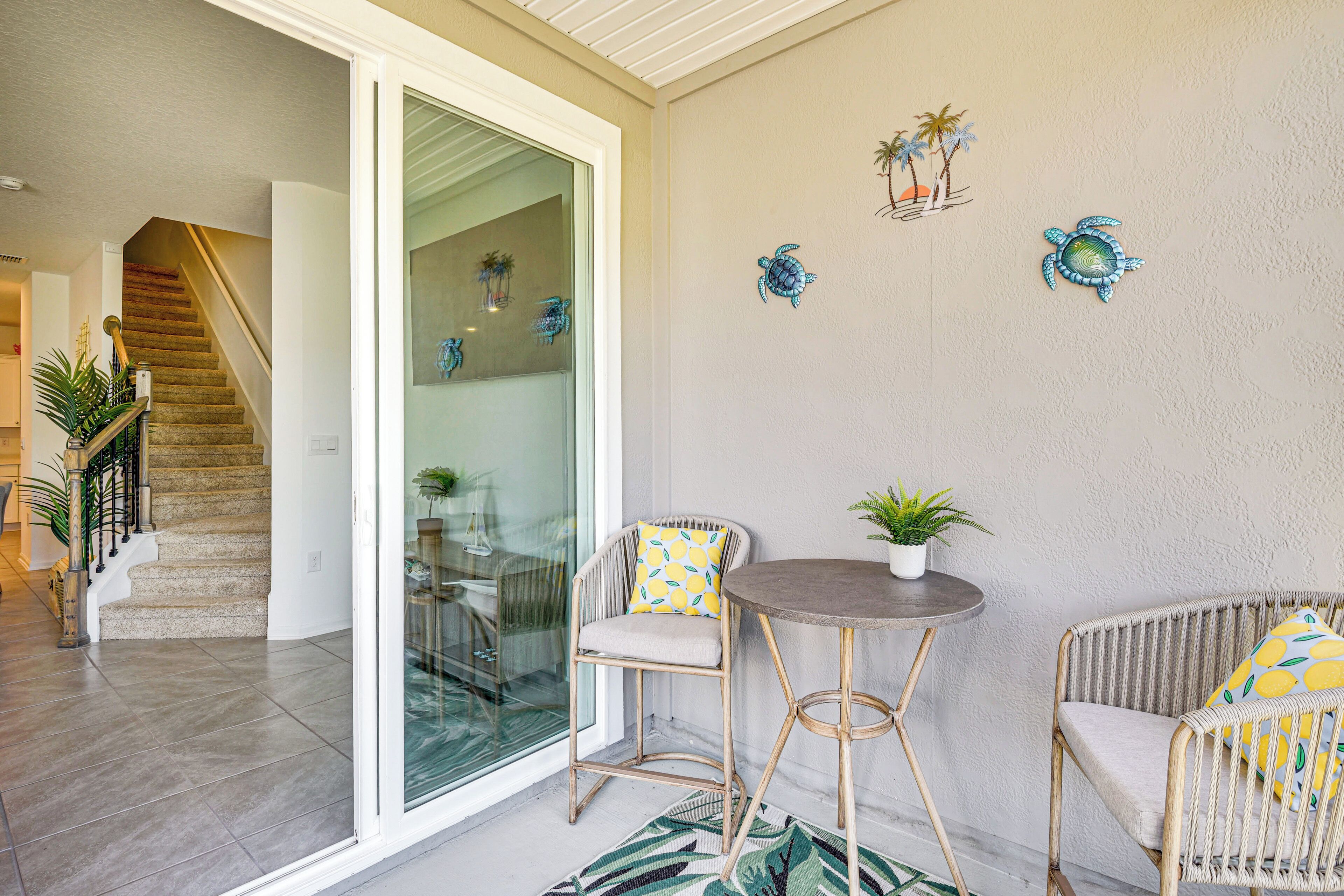 A spacious balcony with a glass door leading to a living area, a round table, wicker chairs, and decorative wall art.