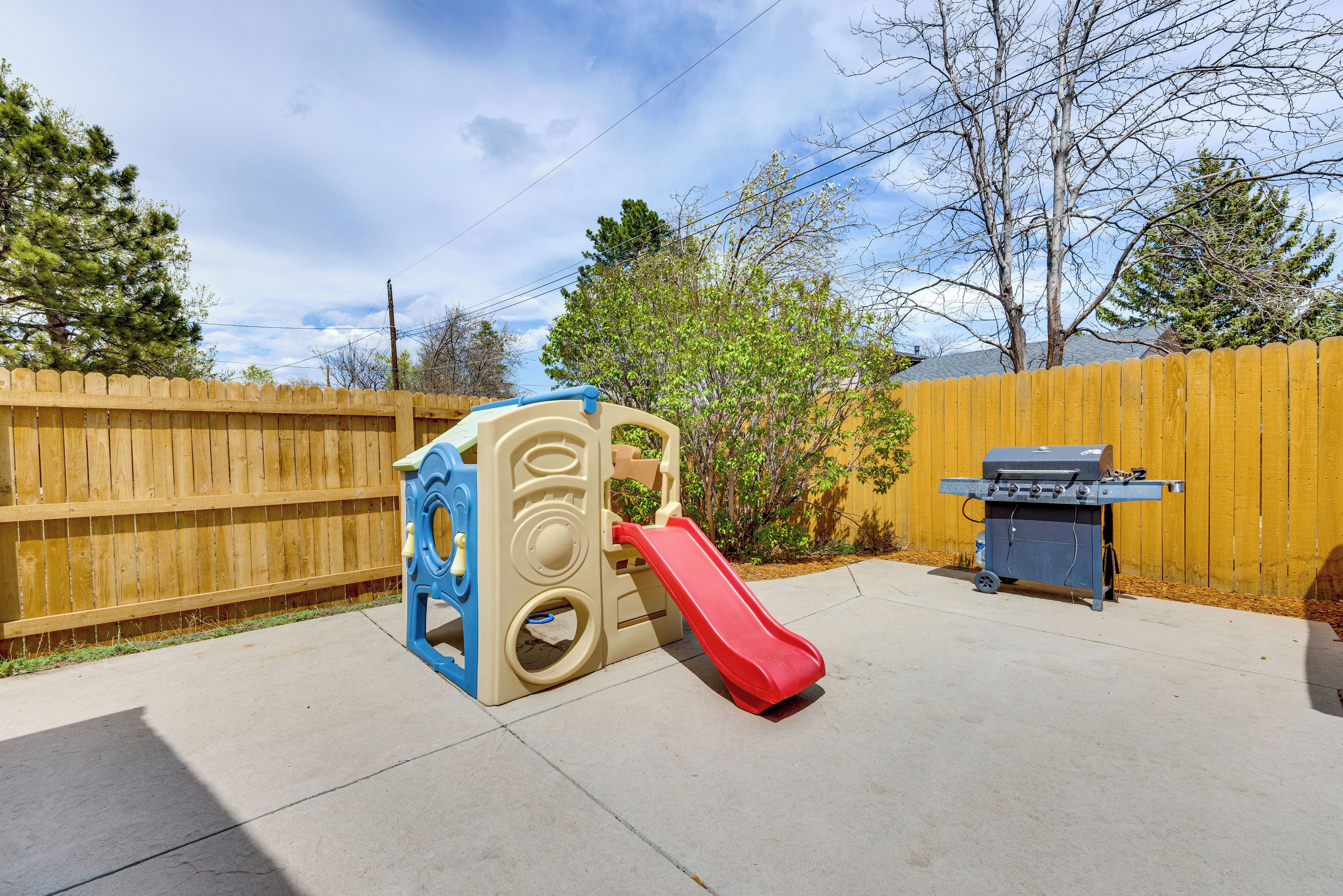 A backyard with a wooden fence, a play structure with a slide, and a barbecue grill.