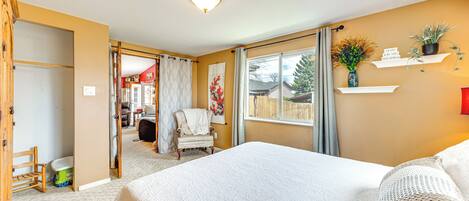 A bedroom with a large bed, a chair, a window with curtains, a shelf with plants, and a door leading to another room.