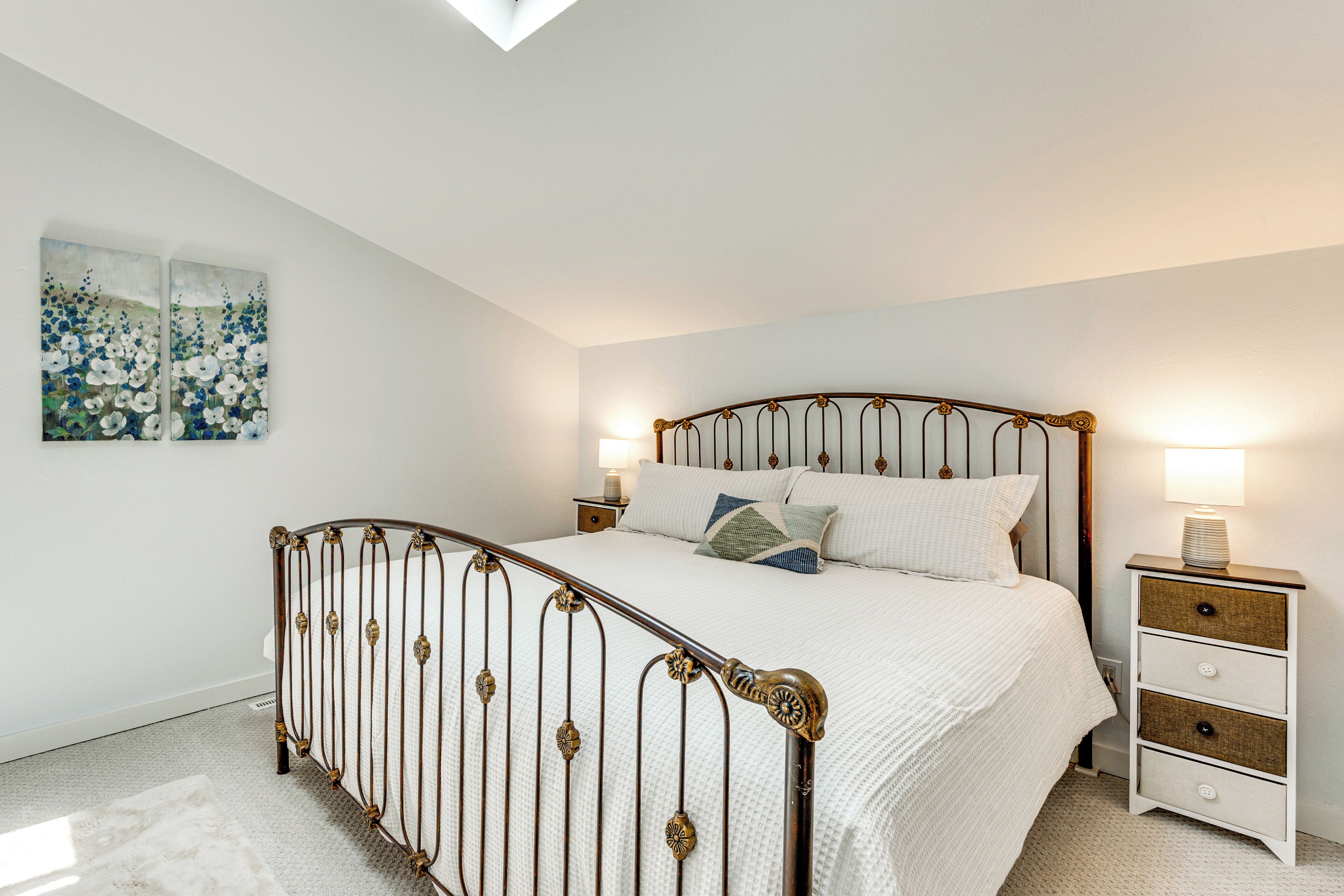 A bedroom with a metal-framed bed, white bedding, a nightstand with a lamp, and a wall-mounted painting.