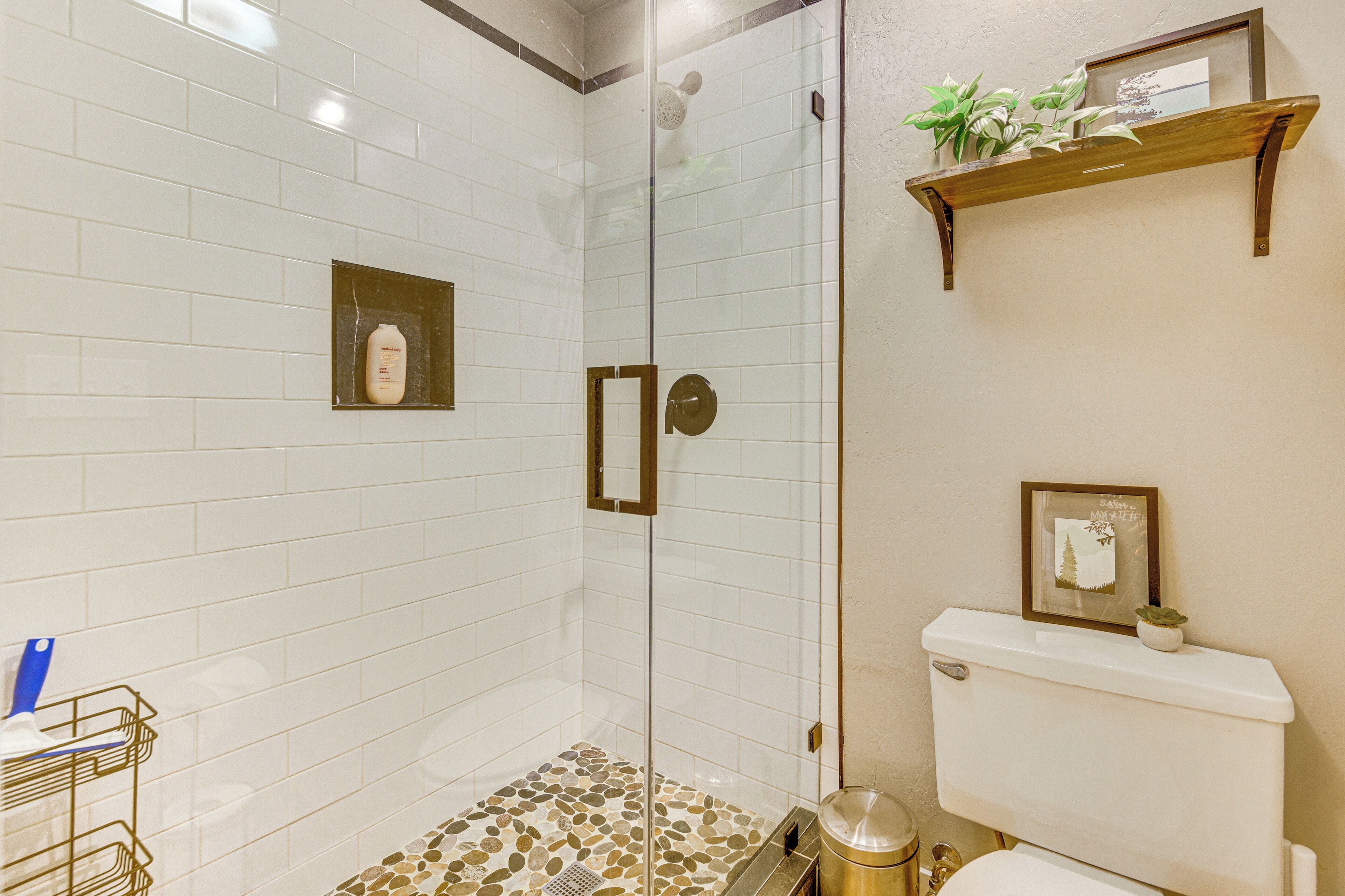 A bathroom with a glass-enclosed shower, white tiled walls, a wooden shelf with a plant, and a toilet with a framed picture.