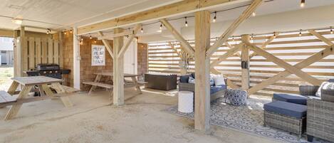 An outdoor covered area with wooden beams, string lights, a barbecue grill, picnic tables, and seating.