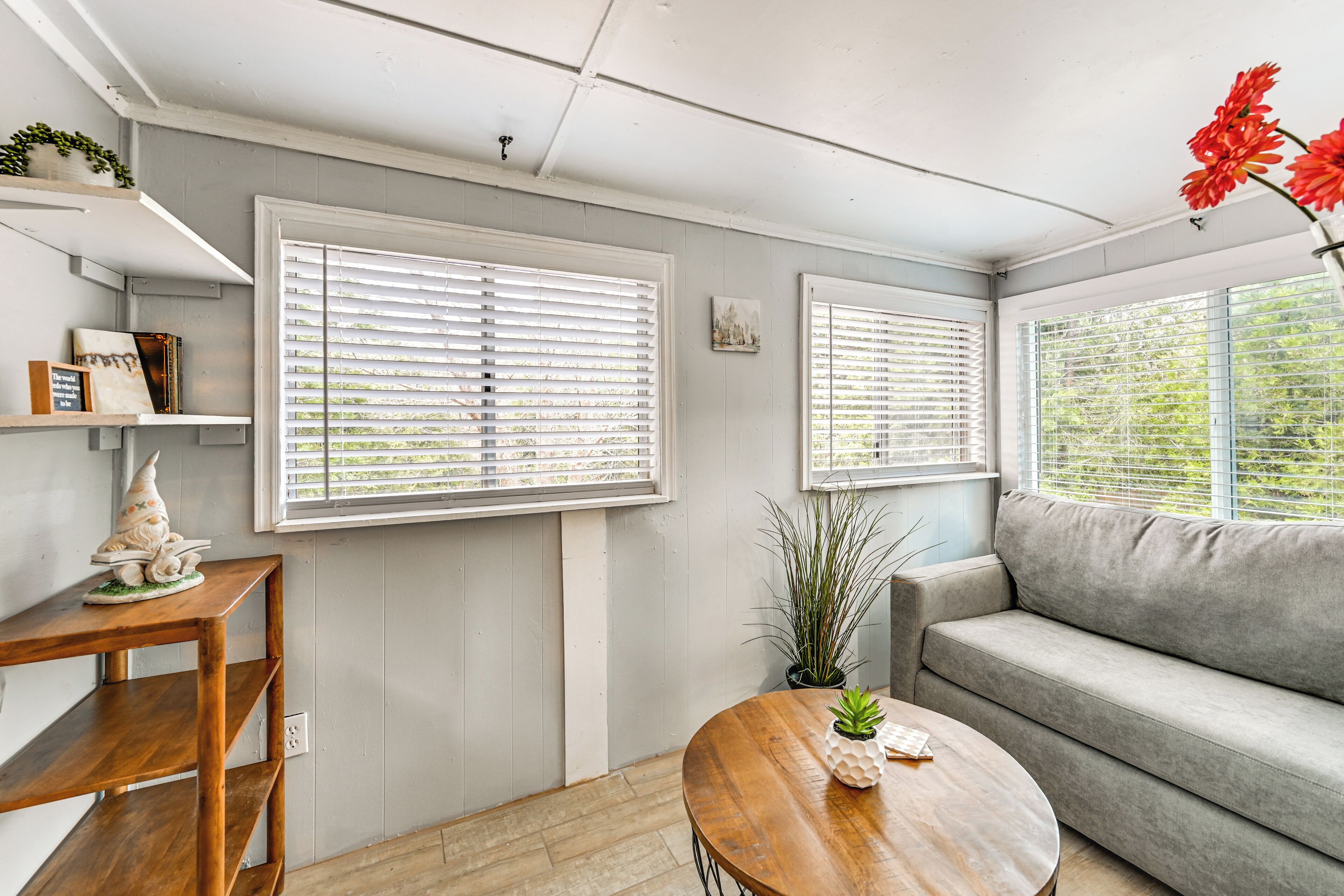 A cozy living room with a wooden coffee table, grey sofa, and white blinds on the windows.