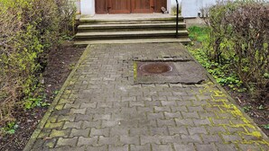 A building entrance with a wooden door, a blue sign above the door, and a paved pathway leading to the entrance.