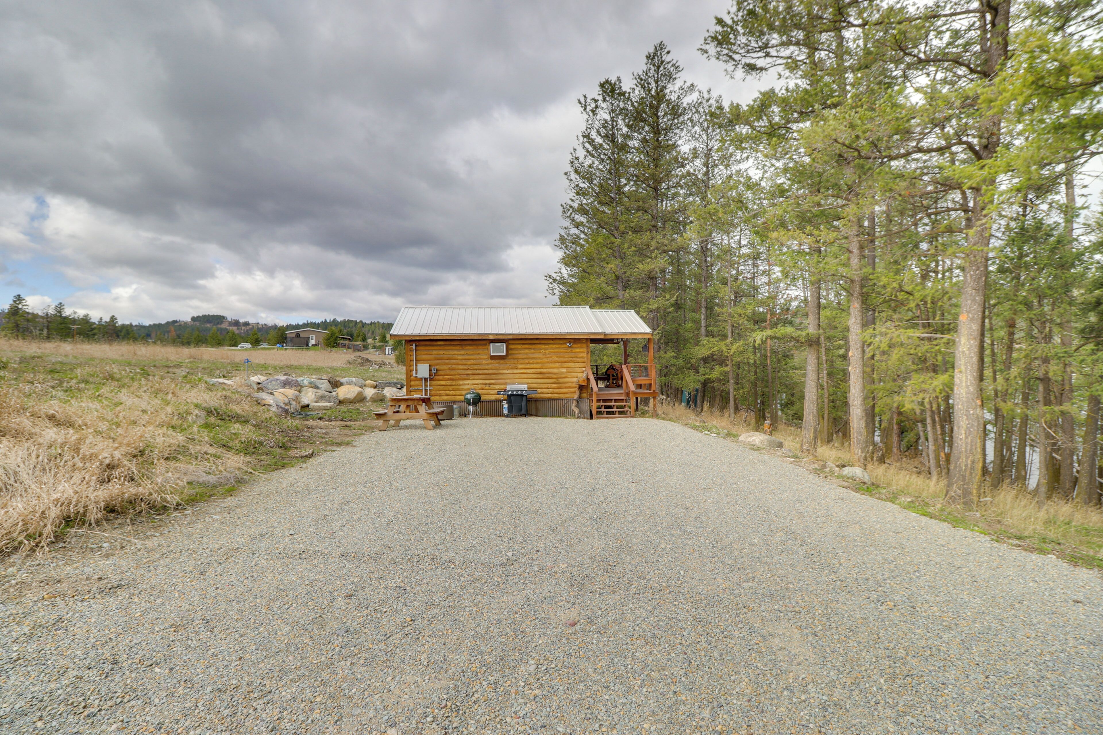 Une cabane en bois avec une allée en gravier, entourée d’arbres et sous un ciel nuageux.