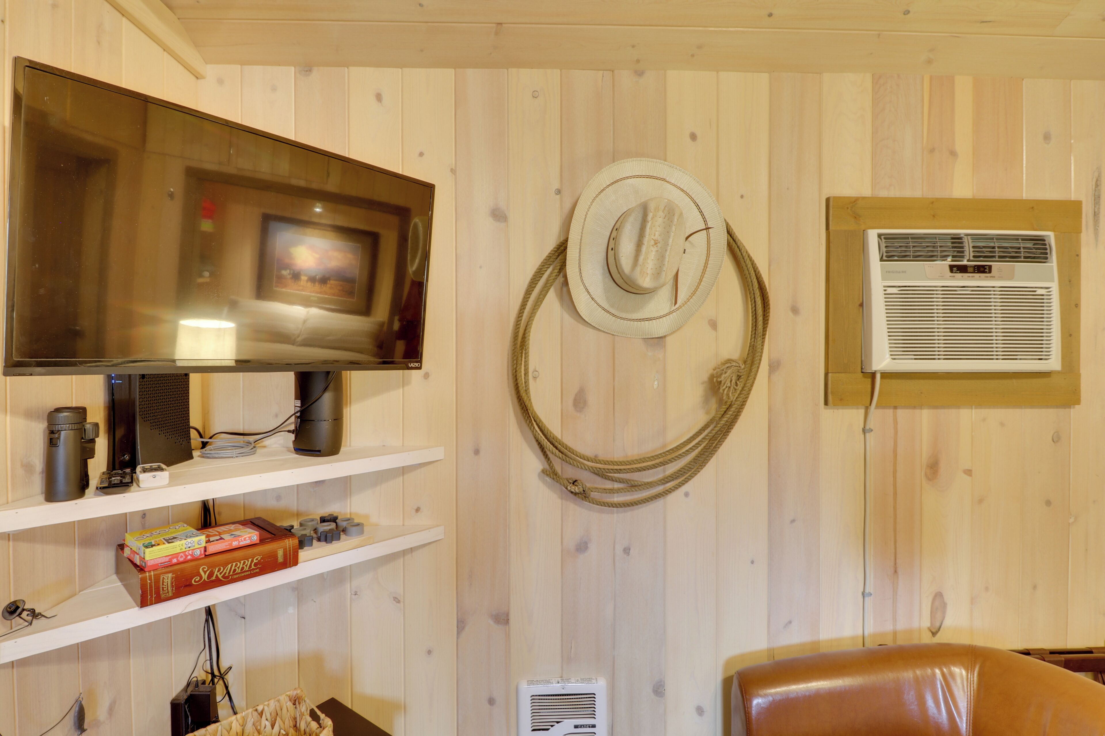 A room with a mounted television, a wall-mounted air conditioner, and shelves with various items including a Scrabble box.