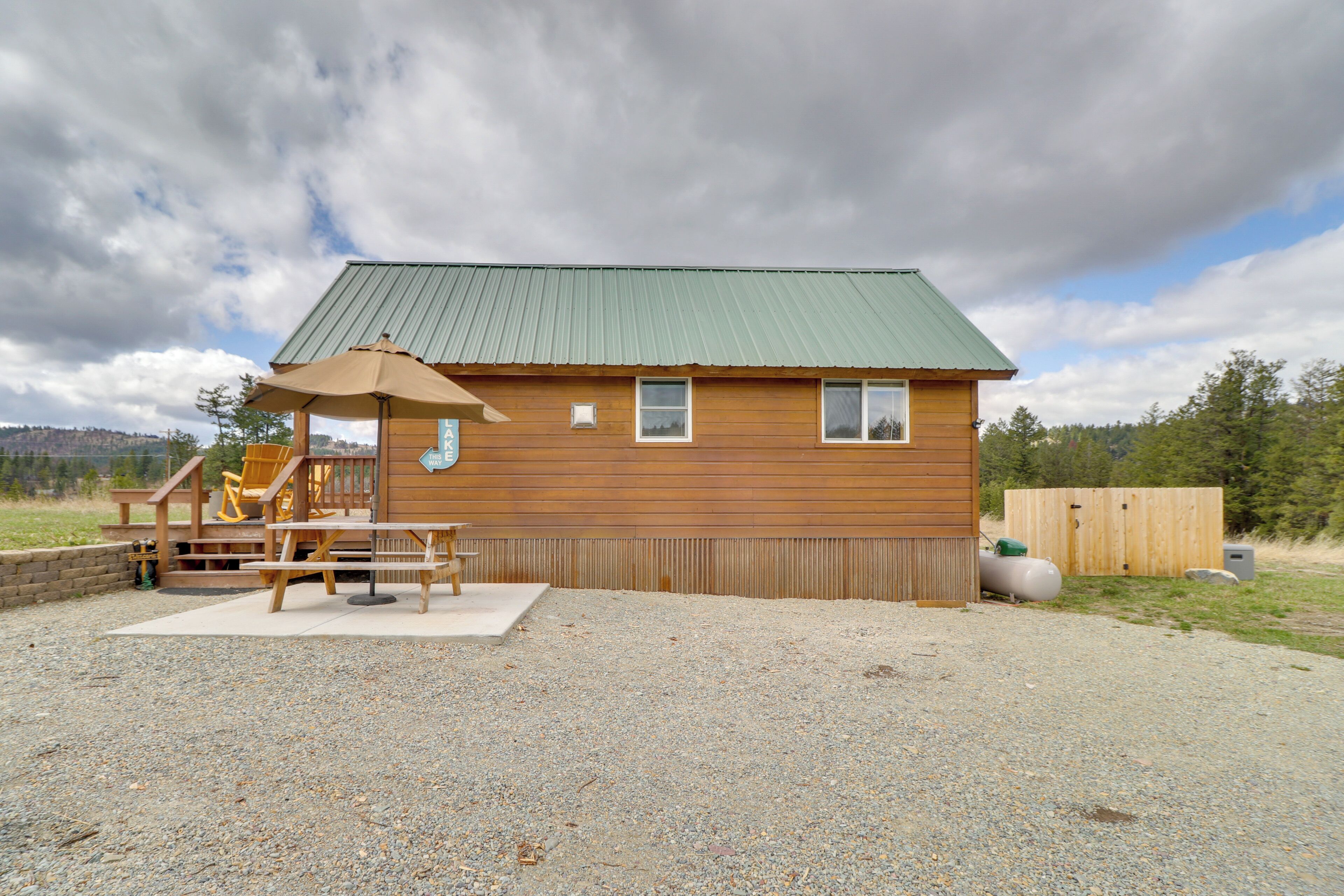 A wooden cabin with a green roof, a covered deck with a table and chairs, and a small staircase leading to the entrance.