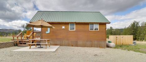 A wooden cabin with a green roof, a covered deck with a table and chairs, and a small staircase leading to the entrance.