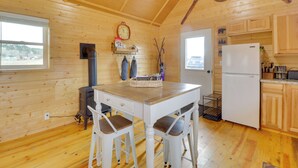 Interior de una cabaña de madera con cocina, una mesa de comedor con dos sillas, un reloj y una ventana.