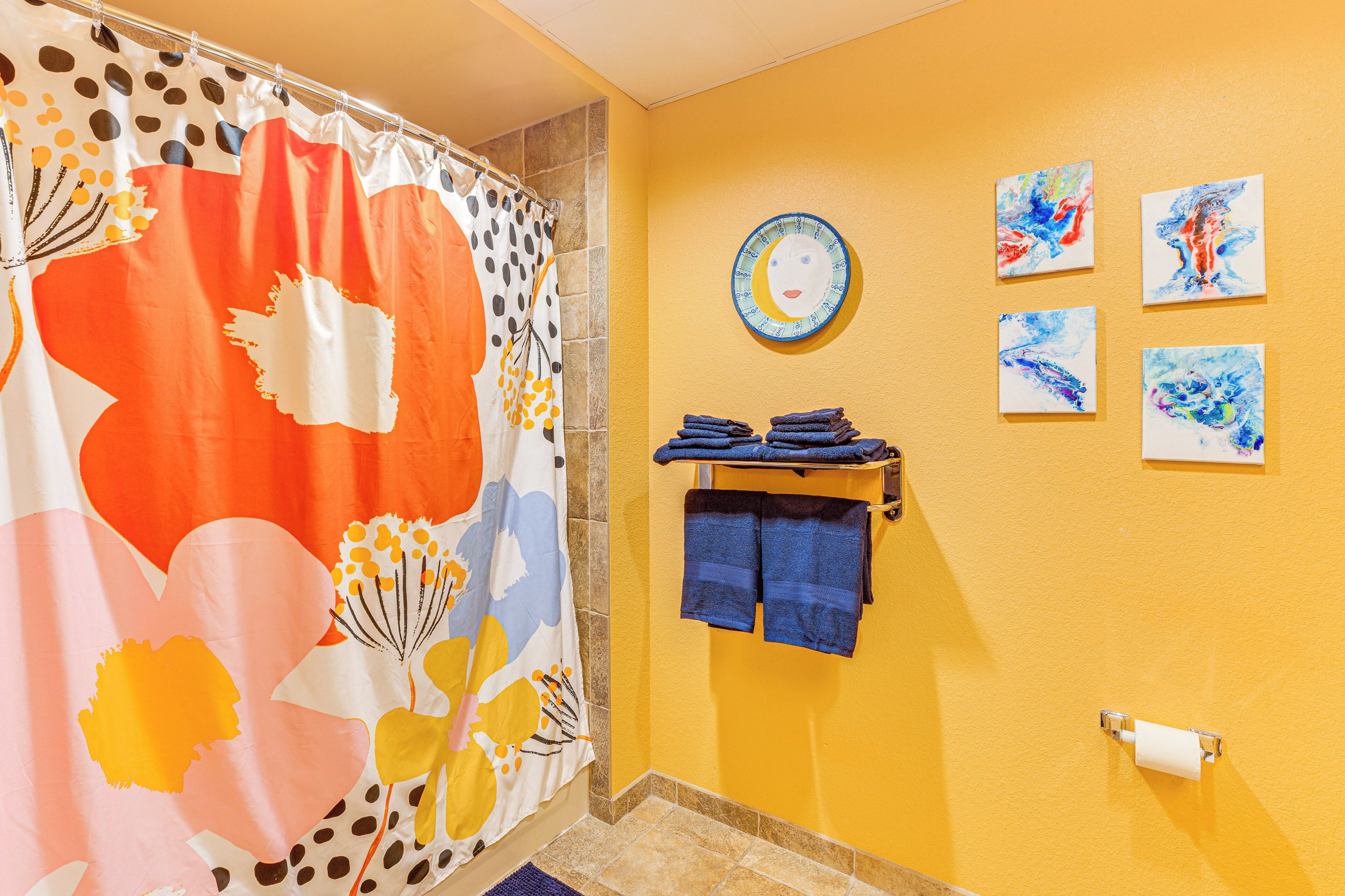 A bathroom with a shower curtain featuring floral patterns, a towel rack with folded towels, and four wall-mounted artworks.