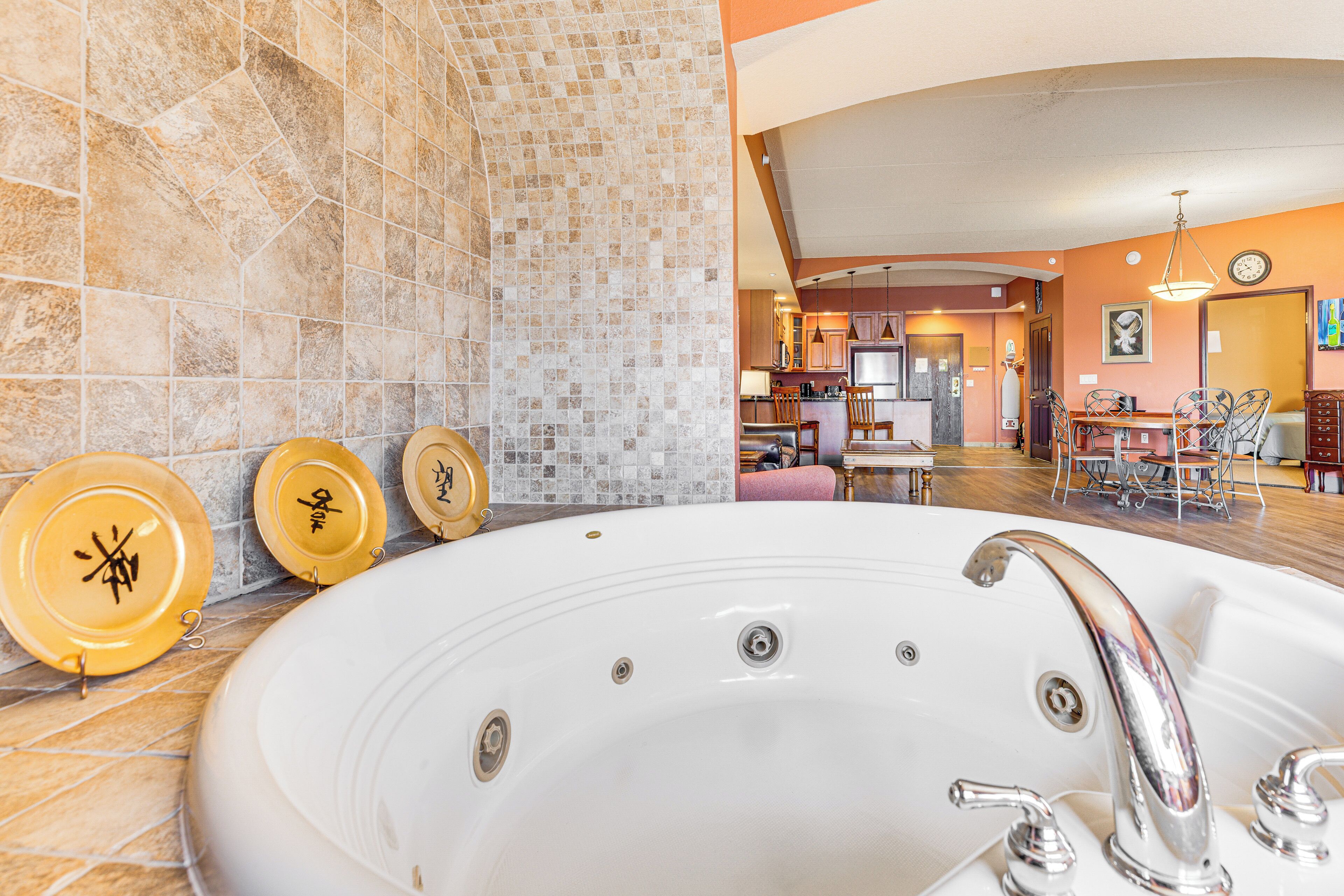 Eine große, weiße Badewanne mit Chromarmaturen, eine mit Mosaik verzierte Wand und im Hintergrund ein Essbereich mit Stühlen und Tisch.