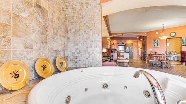 Une grande baignoire blanche avec des robinetteries chromées, un mur carrelé orné d’un motif en mosaïque, et un coin repas avec des chaises et une table en arrière-plan.