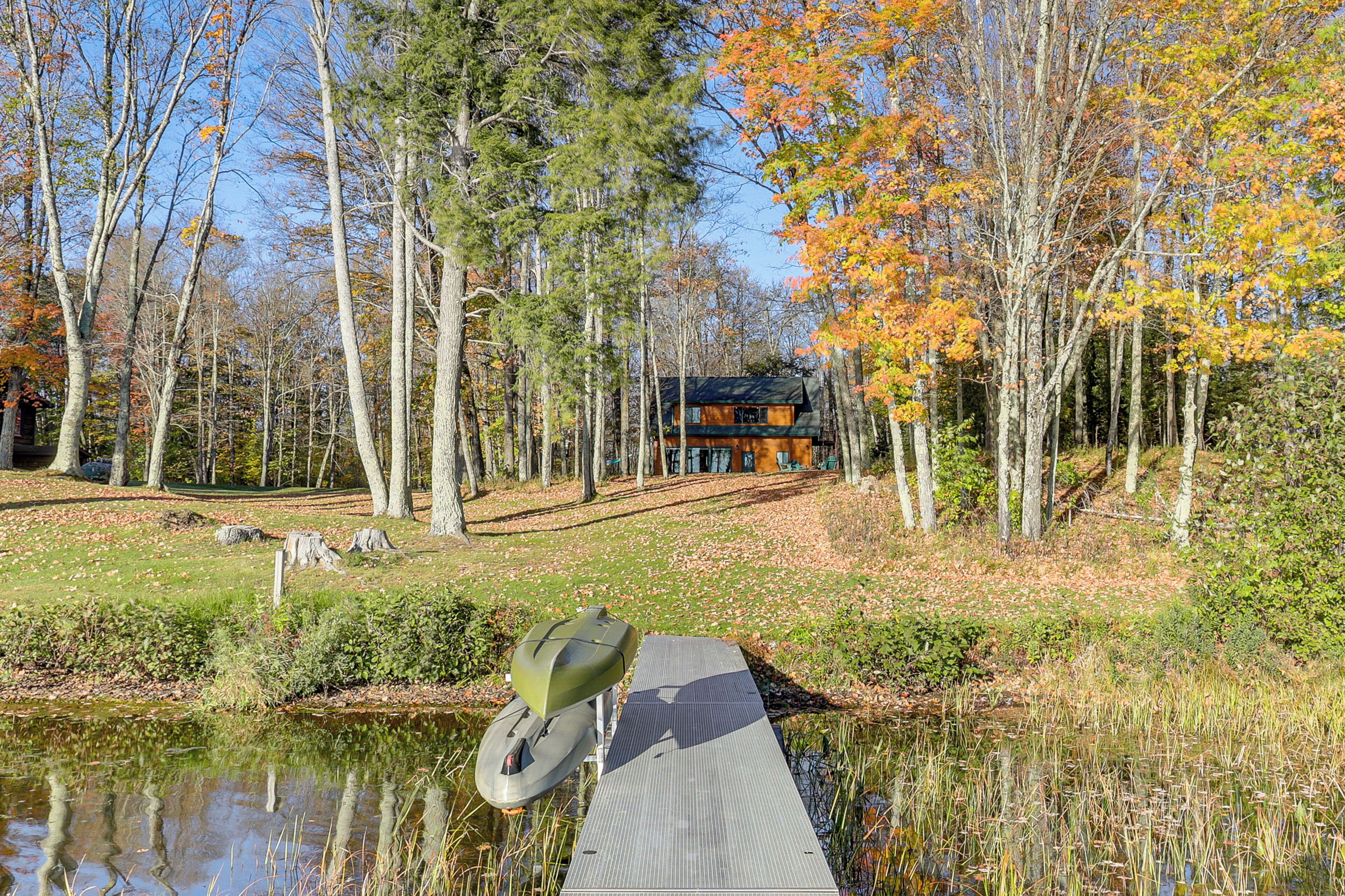 A cabin surrounded by trees with autumn foliage, a dock extending into a pond, and a clear sky.