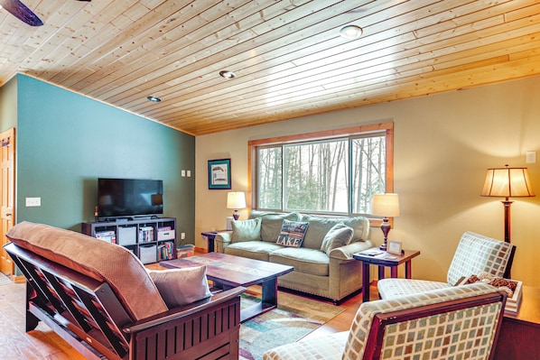 A cozy living room with a wooden ceiling, a television, a sofa, a coffee table, and a window with a view of trees. - Private Dock, BBQ Ready: Lakefront Fifield Home! (Park Falls)