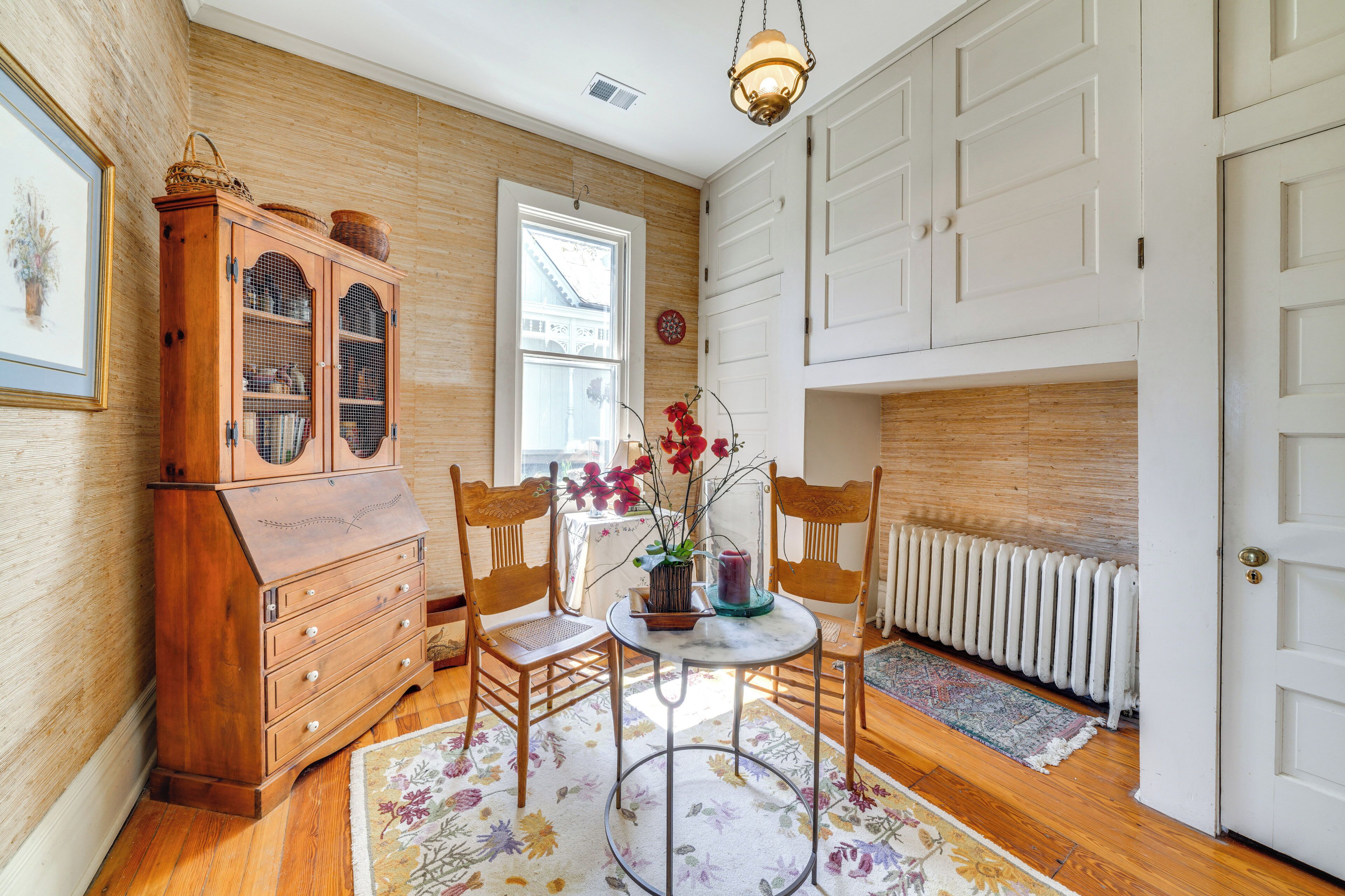 A room with wooden flooring, a wooden cabinet, two chairs, a table with a vase of flowers, a radiator, and a door.