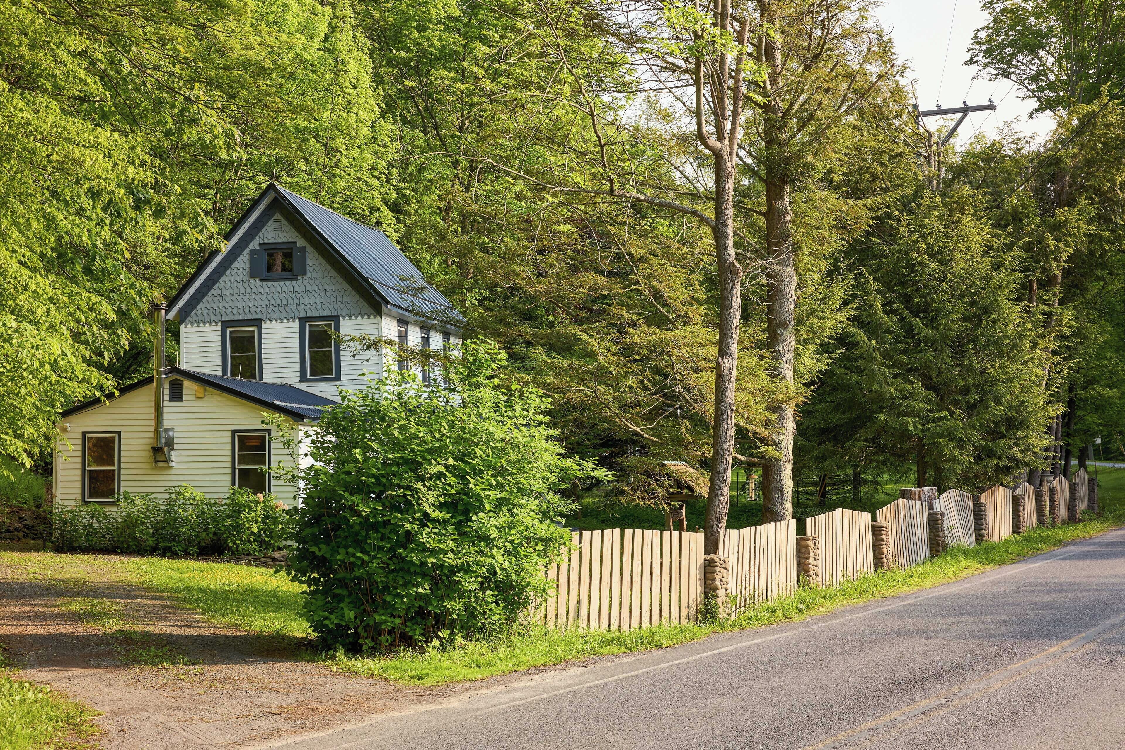 Een huis met twee verdiepingen en een puntig dak, omgeven door dichte bomen en een houten hek.