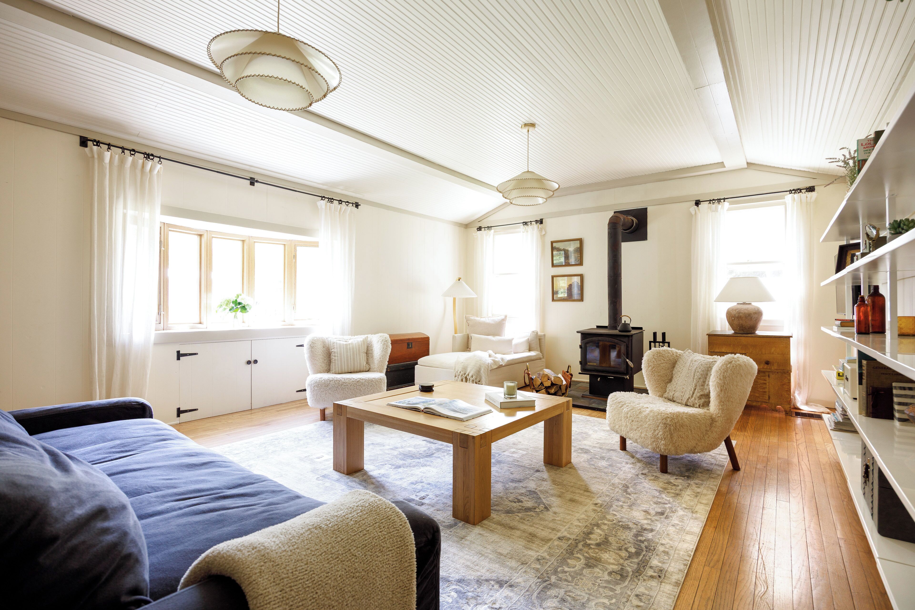 A spacious living room with a sofa, armchair, and coffee table. There is a fireplace, a bookshelf, and a window with curtains.