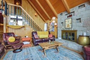 A spacious living area with a wooden staircase, leather furniture, a stone fireplace, and a large rug.