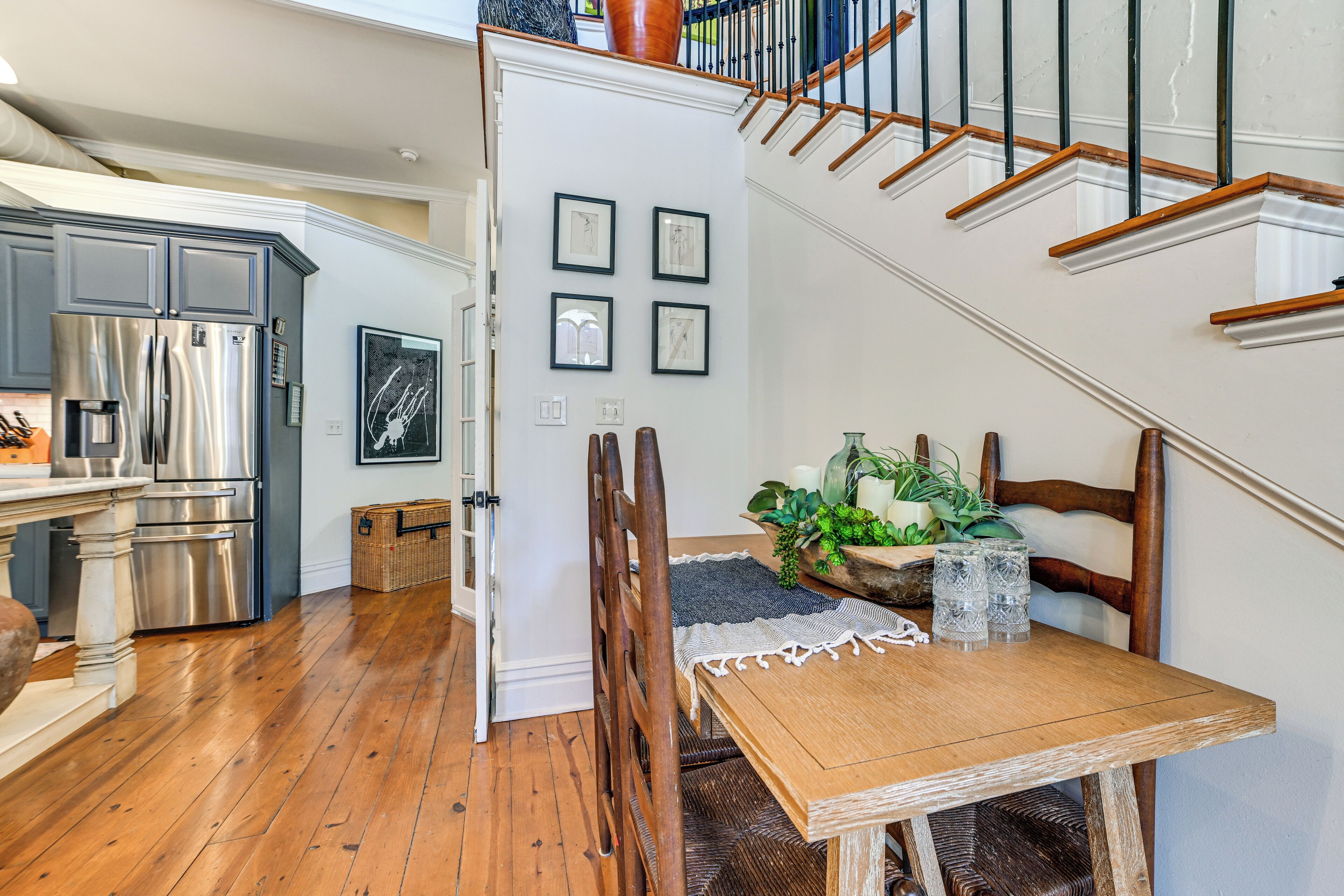 A kitchen with a stainless steel refrigerator, wooden dining table, and chairs. There is a staircase with wooden railings leading to an upper level.
