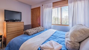A bedroom with a bed, a TV on a dresser, a window with curtains, and a door. - On Mountain two bedroom condo (Newry)