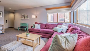 A living room with a large pink sectional sofa, a wooden coffee table, and a teal chair. There is a window with white curtains and a view of a building outside. - On Mountain two bedroom condo (Newry)