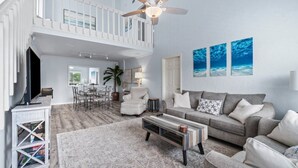 A spacious living room with a high ceiling, a ceiling fan, a grey sofa, a coffee table, and a dining area with a table and chairs.