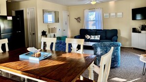 A dining area with a wooden table, white chairs, and a blue ottoman. There is a black refrigerator and a white cabinet in the background. - CH108A- CF w/ 3 Bedrooms, Private Dock, Fire Pit and Dog Friendly (Kill Devil Hills)