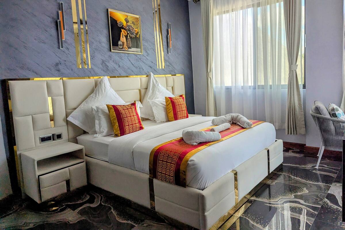 A modern hotel room with a large bed, a nightstand, a bedside table, and a window with sheer curtains.