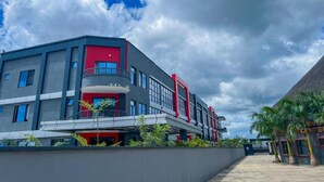A modern building with a red and grey facade, a covered walkway, and a palm tree-lined pathway. - Twalumba Resort & Hotel (Lusaka)