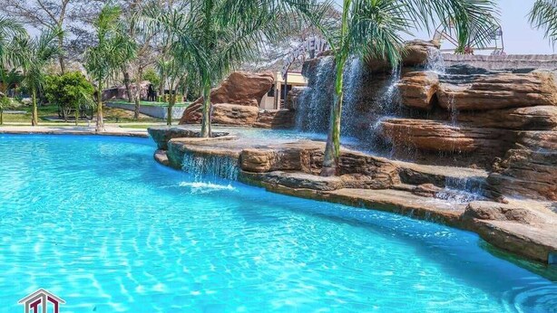 A clear blue pool with a waterfall feature, surrounded by palm trees and rocks. - Twalumba Resort & Hotel (Lusaka)