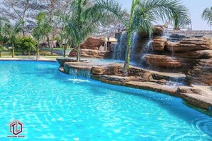 Piscina azul cristalina com cascata, rodeada por palmeiras e pedras.