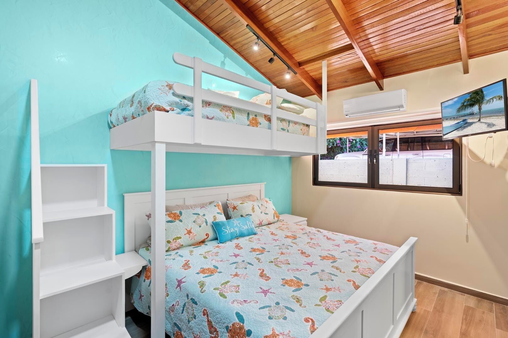 Una cama litera con una cama inferior y otra superior, ambas con ropa de cama estampada. Hay estanterías blancas a la izquierda, una ventana con vistas al exterior y un equipo de aire acondicionado en la pared.