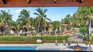 A pool area with a view of a resort complex, including multiple buildings and palm trees. - Stylish 2-Bedroom Beach Condo with Ocean-View Deck, Lush Pool Area & 2-Minute Walk to Flamingo Beach (Cabo Velas)
