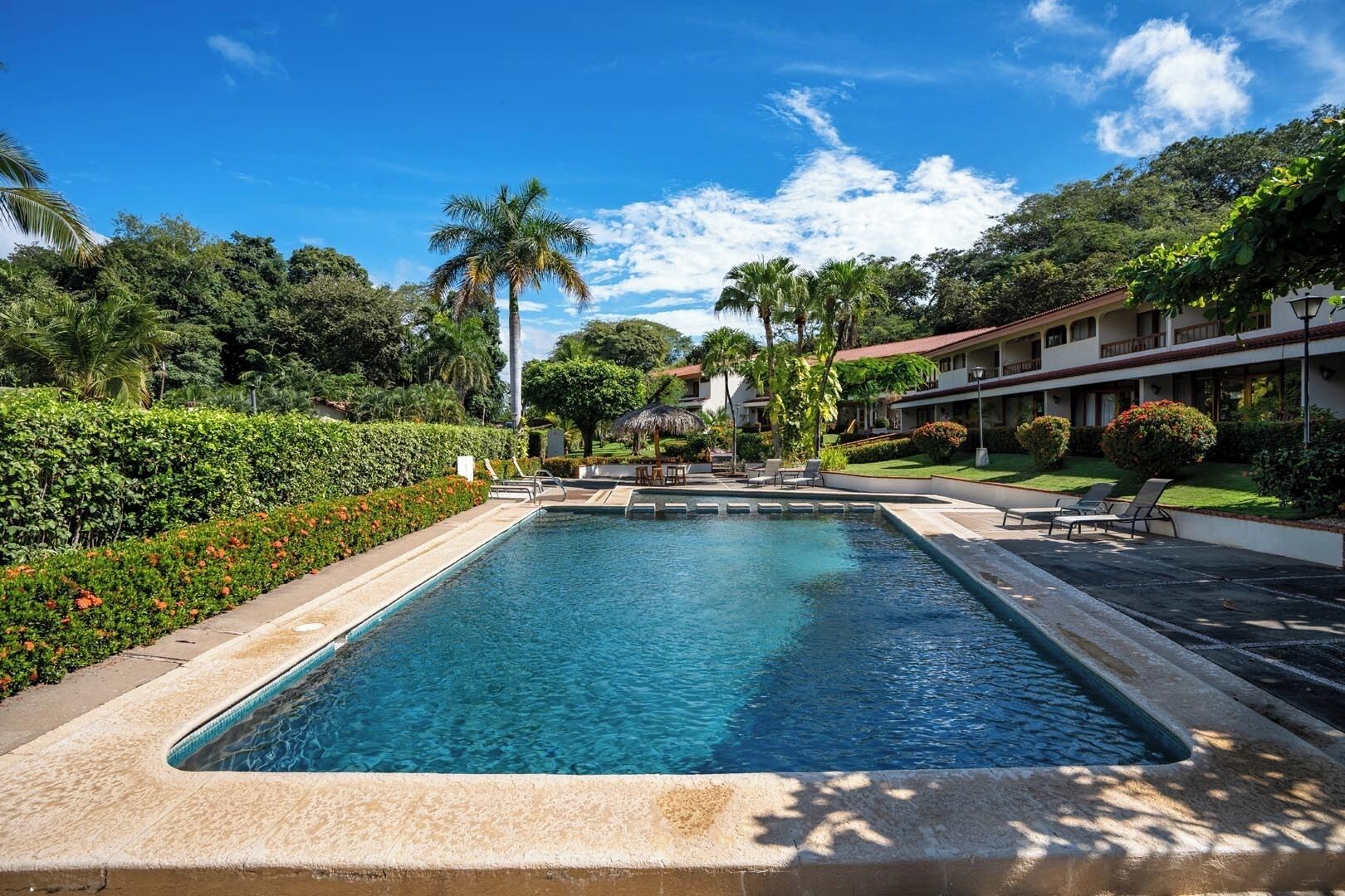 A rectangular swimming pool with a clear blue water surface, surrounded by a low stone wall. There are lounge chairs and a small table on the poolside. The pool is set in a landscaped area with various plants and trees, and a building with a red roof is visible in the background.